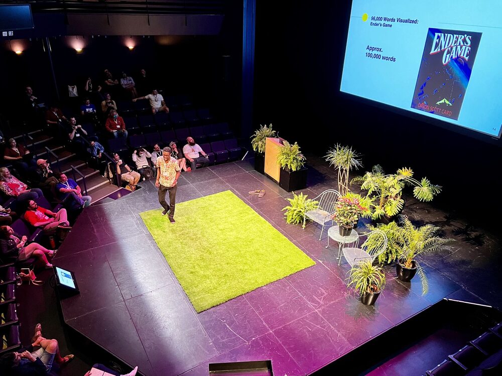 Landon alone on a theatre stage at Madison Ruby, standing on a green astroturf rug among potted plants, a slide behind him visualising a novel's word count