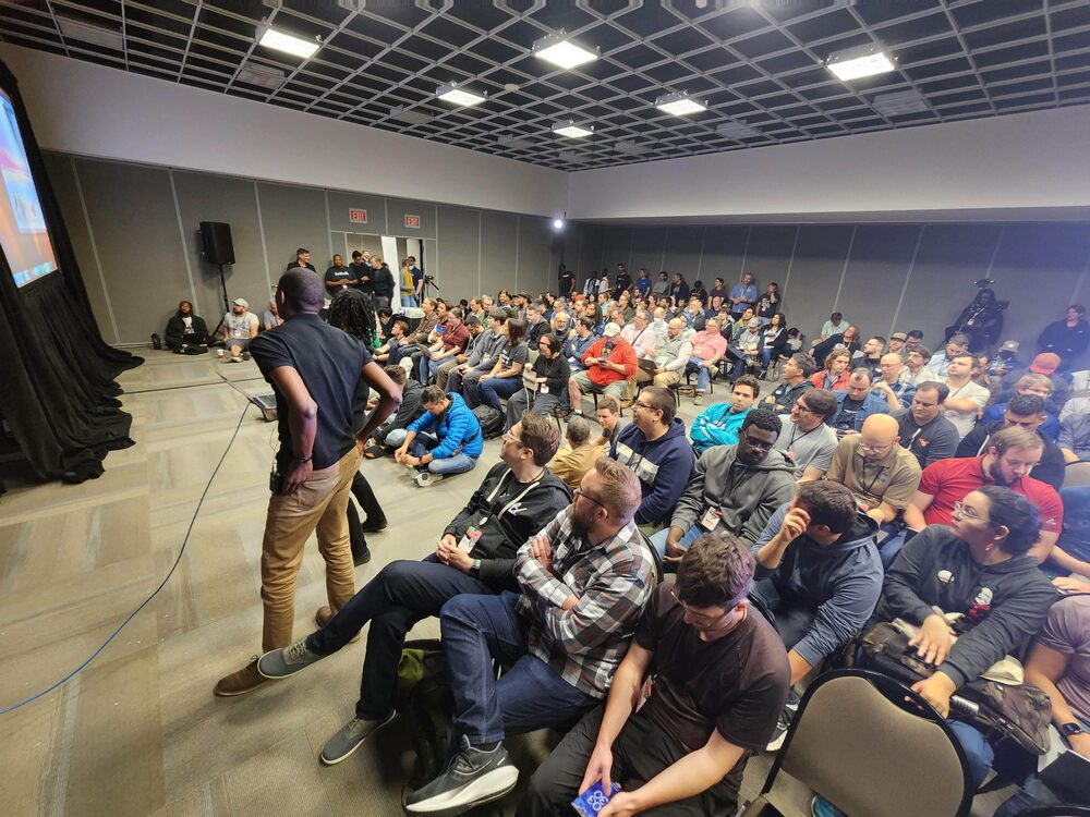 A conference room so full that people are sitting on the floor, Landon speaking at the front